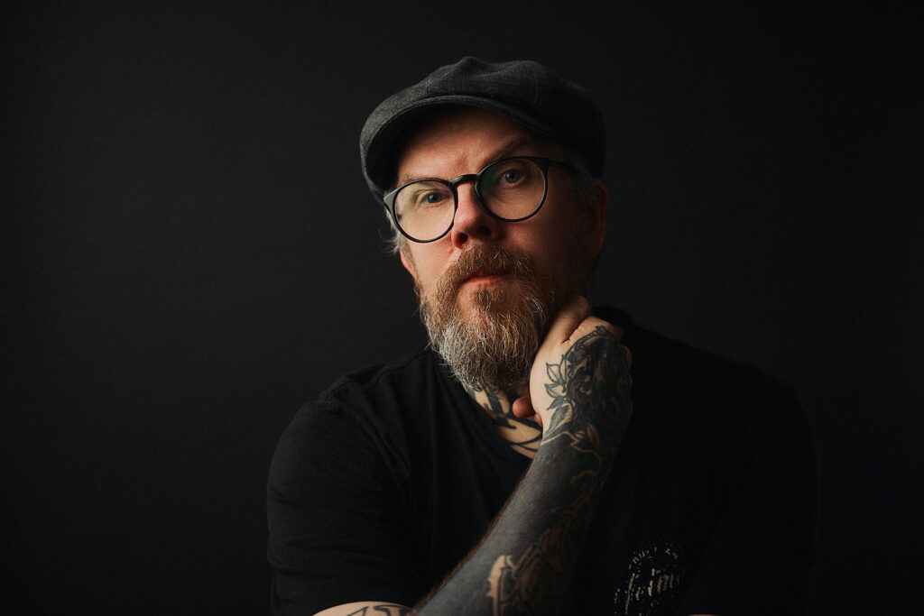 Tatuoija Ilari Musta katsoo kameraan. Hänellä on hattu päässä, musta t-paita, silmälasit. Musta tausta.